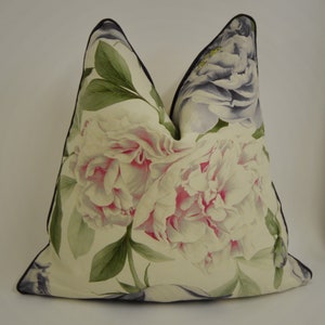 Puede incluir: Un coj&iacute;n decorativo con un gran estampado floral de peon&iacute;as blancas y rosas y hojas verdes. El coj&iacute;n tiene un ribete negro. El patr&oacute;n floral es detallado y realista. El fondo es blanco.