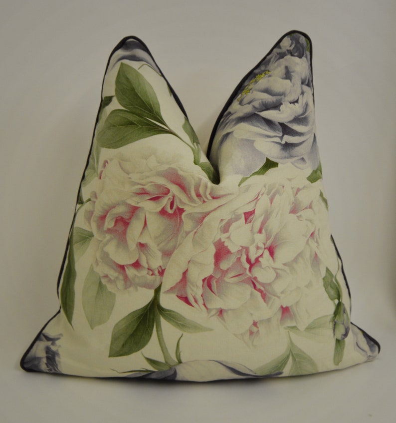 Puede incluir: Un coj&iacute;n decorativo con un gran estampado floral de peon&iacute;as blancas y rosas y hojas verdes. El coj&iacute;n tiene un ribete negro. El patr&oacute;n floral es detallado y realista. El fondo es blanco.