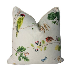 May include: A cream-colored throw pillow with a nature-inspired design featuring green and red birds, a blue butterfly, and green leaves and berries.
