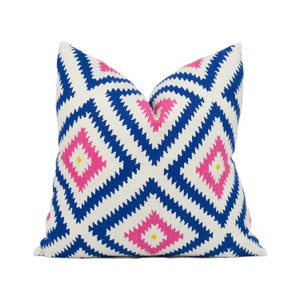 May include: Square decorative pillow with a geometric pattern. The pillow features a white background with a repeating design of blue and pink diamond shapes, each with a small yellow center. The pillow is a square shape.
