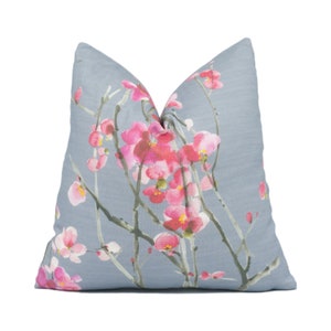 Op de afbeelding: Een vierkant decoratief kussen met een lichtblauwe achtergrond. Het kussen heeft een aquarelontwerp van roze kersenbloesems op grijze takken. Het kussen is een zacht, comfortabel accentstuk voor een bed of bank.