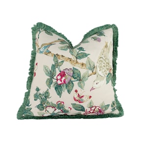 Pode incluir: Uma almofada decorativa com uma borda de franja verde. A almofada apresenta um padrão floral com flores rosa e brancas, folhas verdes e pássaros sobre um fundo creme.