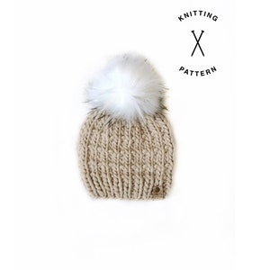May include: A beige knitted beanie with a white faux fur pom pom. The beanie has a ribbed texture and a small brown leather tag with the word "Knitting" on it.