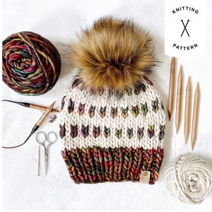May include: A knitted hat with a brown pom pom and a colourful pattern. The hat is made with a white yarn and has a brown, red, and orange stripe at the bottom. The hat is sitting on a white surface with knitting needles and yarn.