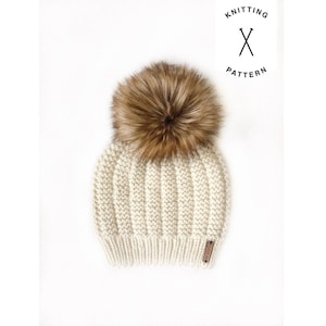 May include: A white knitted beanie with a brown faux fur pom pom. The beanie has a ribbed texture and a small leather label on the side.