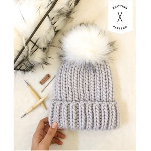 May include: A gray knitted beanie with a white faux fur pom pom. The beanie is made with a chunky yarn and has a ribbed texture. The beanie is being held by a hand.