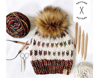 May include: A knitted hat with a brown pom pom and a colorful pattern. The hat is made with a white yarn and has a brown, red, and orange stripe at the bottom. The hat is sitting on a white surface with knitting needles and yarn.