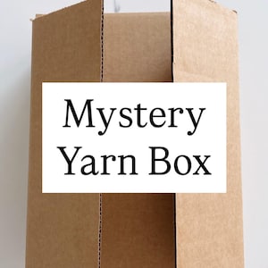 May include: A partially assembled brown cardboard box with the text "Mystery Yarn Box" on a white rectangular label. The box is open, revealing the interior. The background is a white marble surface.