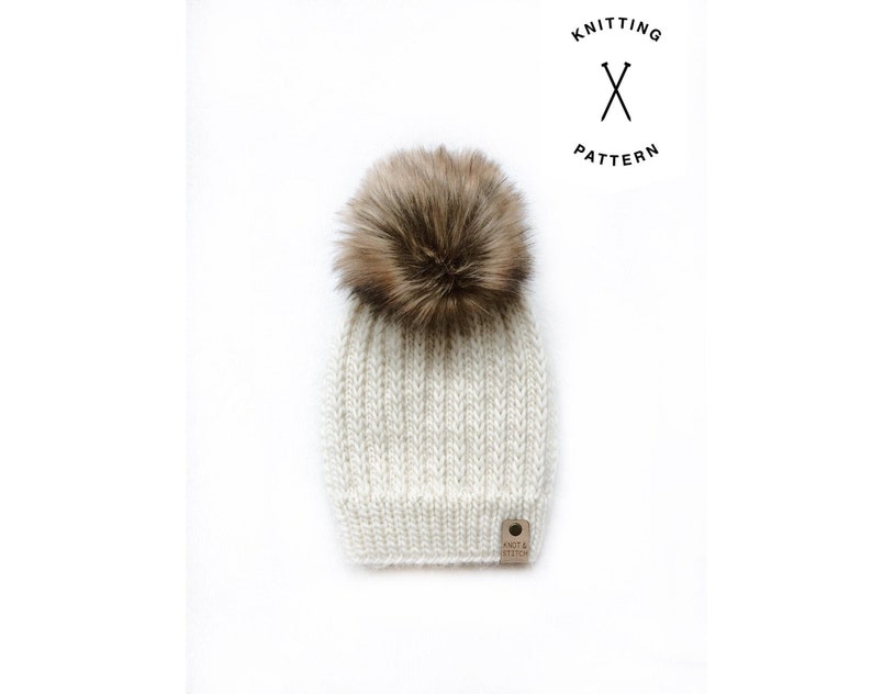 May include: A white knitted beanie with a brown faux fur pom pom. The beanie has a ribbed texture and a small brown leather tag with the text "Knit & Stitch" on it.