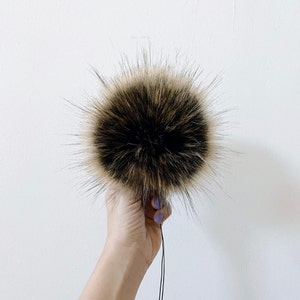 May include: A large, fluffy brown and black faux fur pom pom with a black string attached.