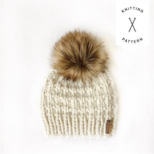 Op de afbeelding: Een witte gebreide beanie met een bruine nep bont pompon. De beanie heeft een textuur gebreid patroon en een klein bruin lederen label met de tekst "Knit & Willow" erop.
