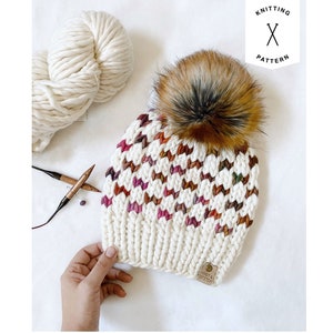May include: A white knitted hat with a colorful chevron pattern and a brown faux fur pom pom. The hat has a brown leather tag with the text "Knit & Stitch" on it.