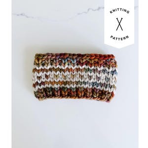 Op de afbeelding: Een dikke, handgebreide hoofdband met een veelkleurig garenpatroon in bruin-, oranje-, wit- en blauwtinten. Het rechthoekige accessoire heeft een grove textuur en bevat een wit label met de tekst KNITTING PATTERN en een icoon van twee gekruiste breinaalden.