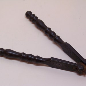 Könnte beinhalten: Ein antiker, schwarzer, hölzerner Nussknacker mit einem Metall-Knackmechanismus. Der Nussknacker hat zwei Griffe mit abgerundeten Enden.