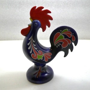 May include: A blue ceramic rooster figurine with red, green, and white painted details. The rooster has a red comb and wattle, and its tail feathers are spread out.