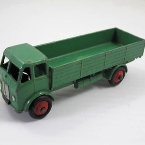 May include: A vintage green toy truck with a cargo bed and red wheels. The truck has a detailed cab with windows and a front grill. The toy truck is made of metal and shows signs of wear, suggesting it is an antique.