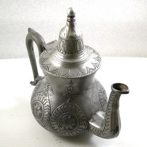 Può includere: Una teiera color argento con intricati disegni incisi. La teiera presenta un manico curvo, un beccuccio con un motivo decorativo e un coperchio a cupola con un pomello. Il corpo della teiera è adornato con motivi floreali e a stella.