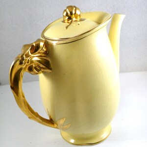 Peut inclure: Une cafetière en céramique jaune avec une poignée et un couvercle dorés. La poignée est en forme de feuille stylisée et le couvercle a un bouton doré.