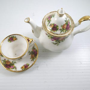 Puede incluir: Un juego de té de porcelana blanca con un diseño floral. El juego incluye una tetera, una taza de té y un platillo. Los bordes están adornados con oro. El diseño floral presenta rosas rosas, amarillas y moradas.