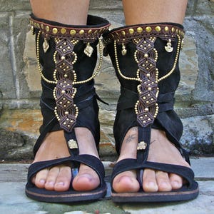 May include: Black leather sandals with gold accents and intricate braided straps. The sandals have a high ankle strap and a t-strap design.