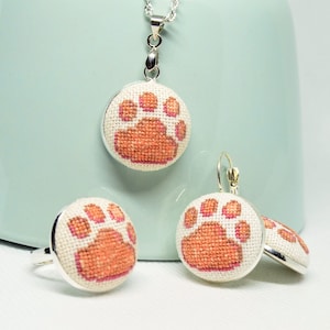 May include: A set of jewelry featuring a necklace, earrings, and a ring. Each piece has a white background with a pink paw print design.