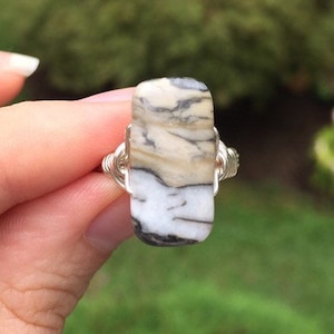 May include: A rectangular ring featuring a natural stone with a mix of white, beige, and black patterns. The stone is set in a silver-colored wire wrap design, creating a unique and earthy aesthetic.