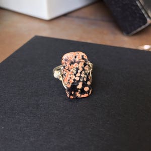 May include: A silver wire wrapped ring featuring a rectangular, black and orange speckled stone.