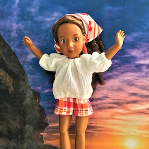 May include: A doll with brown hair and brown eyes wearing a white blouse, red and white checked shorts, and a red and white patterned headscarf. The doll is standing on a rock with a sunset and ocean in the background.