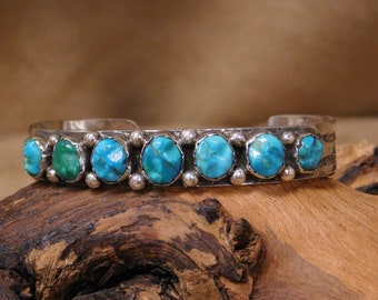 May include: A silver cuff bracelet featuring seven oval turquoise stones and one green stone. The stones are set in individual bezels and framed by small silver beads. The bracelet rests on a piece of wood.