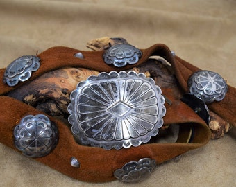 May include: A brown leather belt with silver concho accents. The belt is decorated with several round and oval conchos, each with a unique design. The belt is folded and lying on a brown fabric surface.