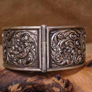 May include: A silver bracelet with intricate floral designs. The bracelet is hinged and opens to fit the wrist.