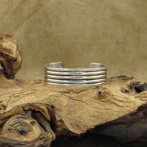 May include: A silver bracelet with five bands, each with a slightly different width. The bracelet is on a wooden surface.