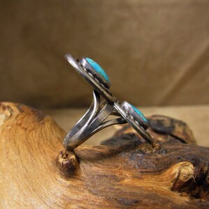 May include: A silver ring with two turquoise stones set in a decorative design. The ring is on a wooden surface.