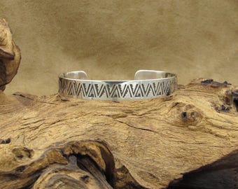 May include: A silver bracelet with a geometric pattern of triangles. The bracelet is on a wooden surface.