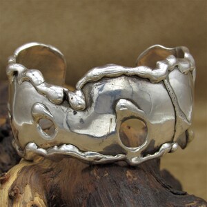 May include: A silver cuff bracelet with an organic, textured design. The bracelet has several open circles throughout the design.