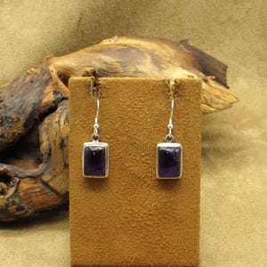 May include: A pair of silver-tone earrings with rectangular, deep purple amethyst stones. The earrings are displayed on a brown suede-like stand against a natural wood background. The earrings have a simple, elegant design.