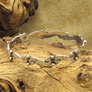 May include: A silver bangle bracelet with a wavy design and small, round accents. The bracelet is displayed on a wooden surface, showcasing its intricate details and craftsmanship. The bracelet's design is a continuous loop.