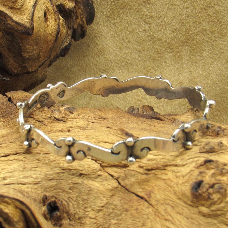 May include: A silver bangle bracelet with a wavy design and small, round accents. The bracelet is displayed on a wooden surface, showcasing its intricate details and craftsmanship. The bracelet's design is a continuous loop.