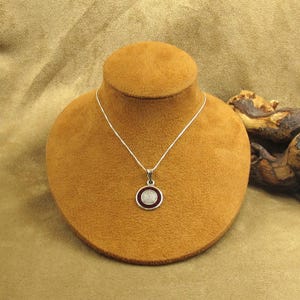 May include: A silver necklace with a circular pendant. The pendant has a dark red center with a silver border. The necklace is displayed on a brown jewelry bust against a brown background. The necklace is a piece of jewelry.