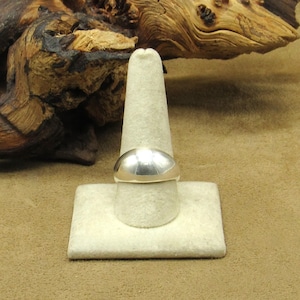 Peut inclure: Une bague en argent poli avec un dessus arrondi est pr&eacute;sent&eacute;e sur un pr&eacute;sentoir blanc. La bague refl&egrave;te la lumi&egrave;re, et l'arri&egrave;re-plan comprend un morceau de bois flott&eacute;. Design simple et &eacute;l&eacute;gant.