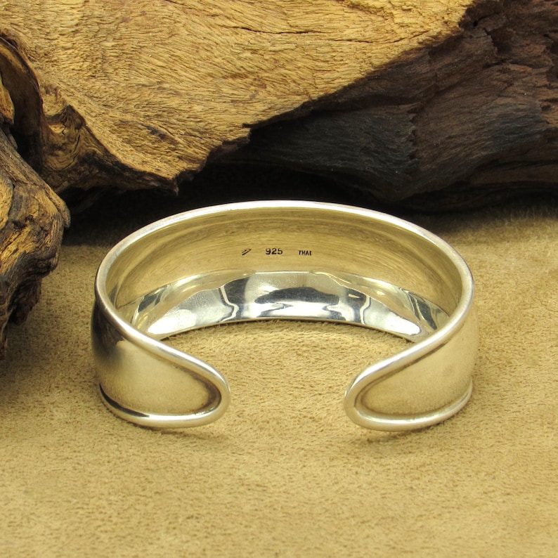 May include: A polished silver cuff bracelet with a curved design. The open ends have rounded edges. The inside of the bracelet is stamped with "925" and "THAILAND". The bracelet is displayed on a tan surface with a wooden background.