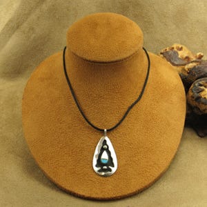 May include: A silver pendant necklace with a black cord. The pendant has a teardrop shape with a turquoise stone and white accents. The necklace is displayed on a brown jewelry bust.