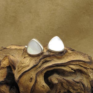 May include: A pair of sterling silver stud earrings. Each earring features a triangular, pearlescent, mother-of-pearl stone set in a silver bezel. The earrings are displayed on a piece of weathered wood.