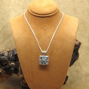 May include: A silver necklace with a square pendant featuring blue gemstones and small white accents. The pendant is suspended from a delicate silver chain. The necklace is displayed on a brown jewelry bust.