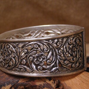 May include: A silver bangle bracelet with intricate floral designs. The bracelet is made of openwork silver and has a hinged clasp.