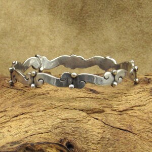 May include: A silver-toned bangle bracelet with a wavy design. The bracelet features a series of curved shapes and small, round embellishments. The metal has a slightly aged appearance, suggesting it may be vintage or antique.