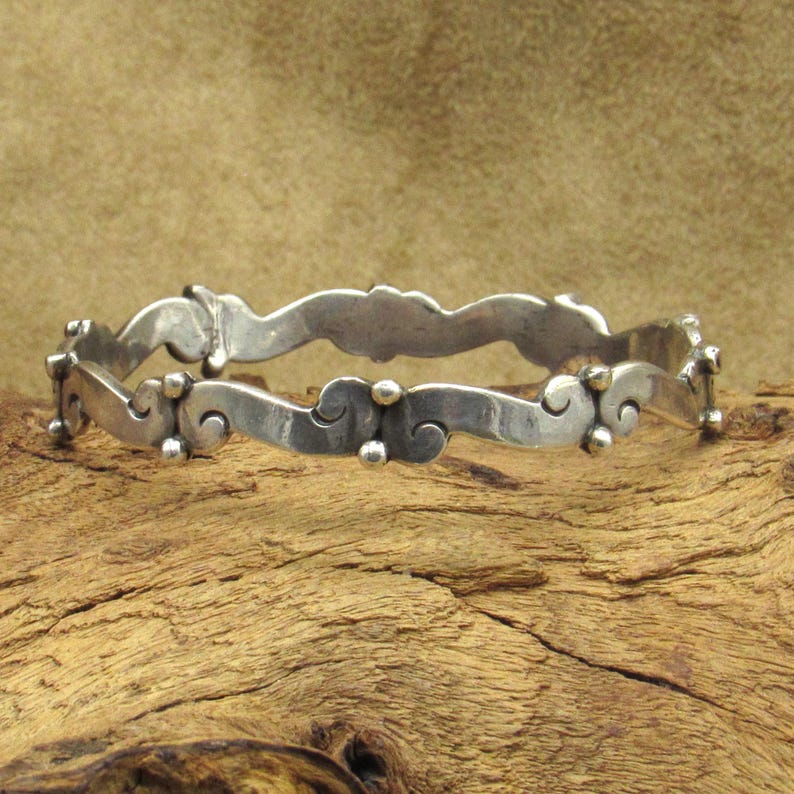 May include: A silver-toned bangle bracelet with a wavy design. The bracelet features a series of curved shapes and small, round embellishments. The metal has a slightly aged appearance, suggesting it may be vintage or antique.
