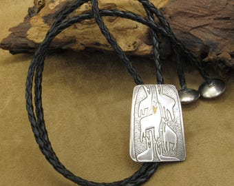 May include: A silver bolo tie with a rectangular pendant featuring a stylized design of a tree with multiple animals. The pendant has a textured surface and a small gold accent. The bolo tie is on a black leather cord.