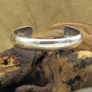 May include: A polished silver cuff bracelet with a rounded design. The bracelet has an open design and is displayed on a piece of wood. The inside of the bracelet has stamped markings. A simple, elegant piece of jewelry.