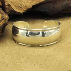 May include: A polished silver cuff bracelet. The bracelet has a wide, rounded band with a smooth, reflective surface. The design is simple and elegant, with a slightly curved shape that tapers towards the opening. The cuff is approximately 2.5 inches wide.
